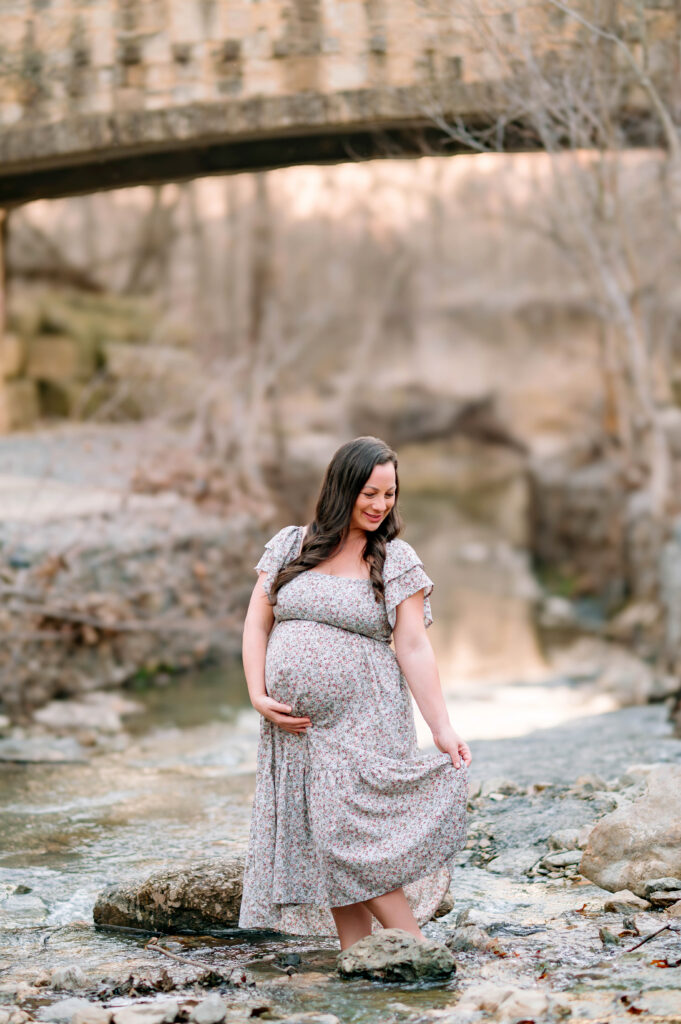 Mom holding pregnant belly and holding dress while looking down and standing in creek during a maternity session at Arbor Hills Nature Preserve in Plano, Texas. 