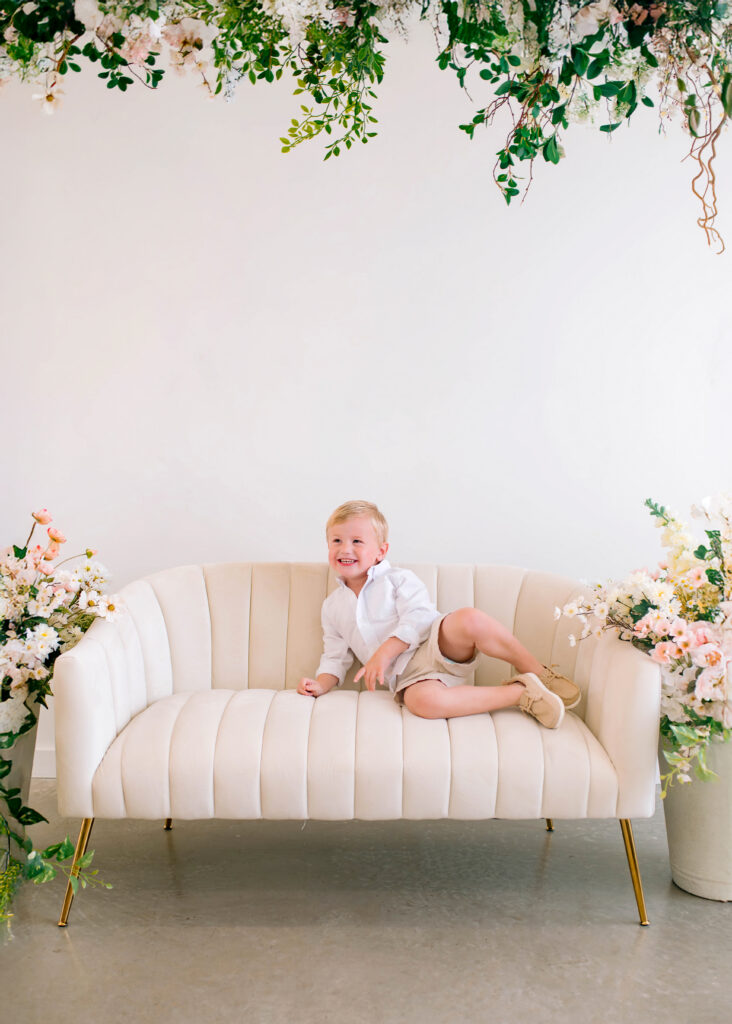 Son laughing and posing on white couch during a spring birth announcement photo session at Lemon Drop Studios in McKinney, Texas.