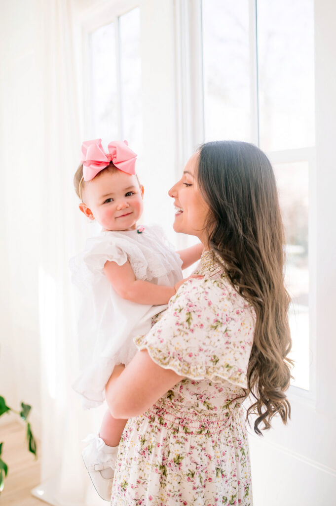 Mom holding daughter front of window with daughter smiling at camera during a portrait session with Jennifer L Kirk photography in Mckinney, Texas. 