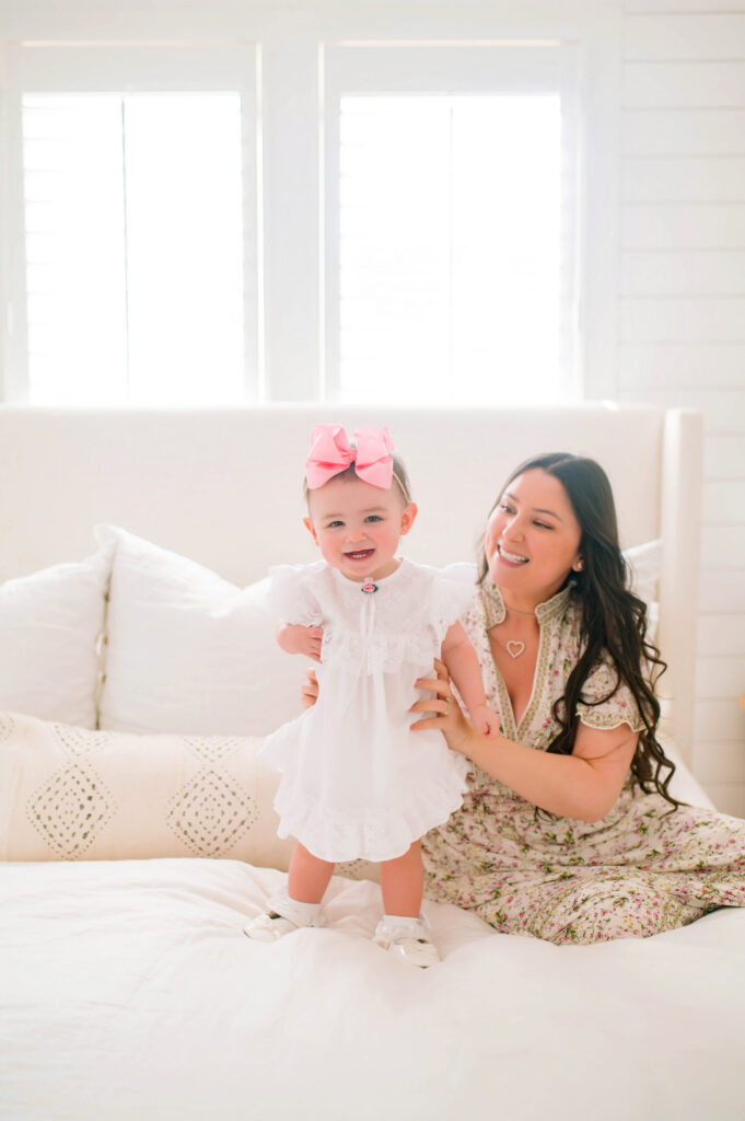 Mom holding daughter standing on white bed during a mommy and me portrait session in Mckinney, Texas. 