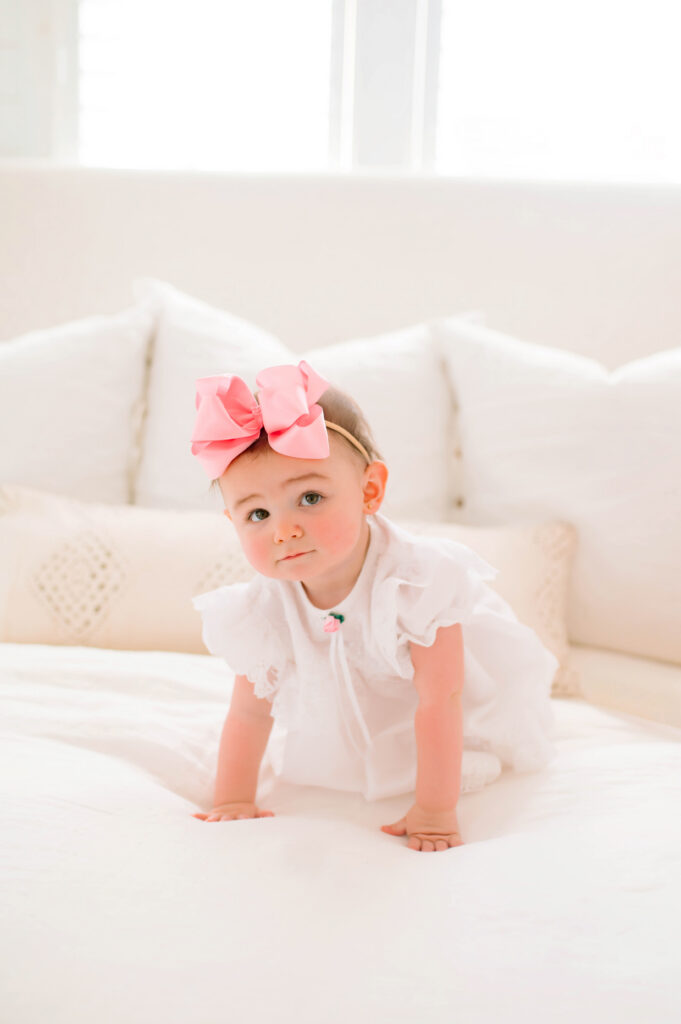 Toddler girl crawling on white bed looking seriously at camera during a portrait session at the Beverly Harris House in Mckinney, Texas. 