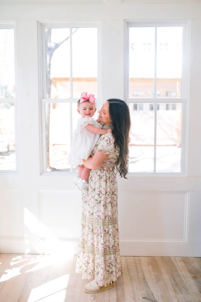Mom and daughter dancing in front of window with daughter smiling at camera during a portrait session with Jennifer L Kirk photography in Mckinney, Texas. 