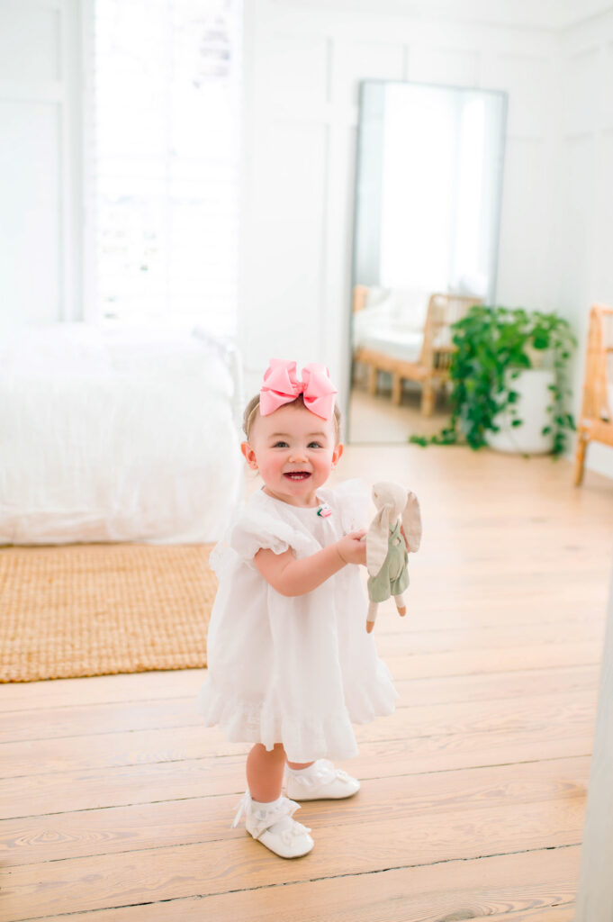 Toddler girl standing in room smiling at camera holding a bunny during a portrait session at the Beverly Harris House in Mckinney, Texas. 