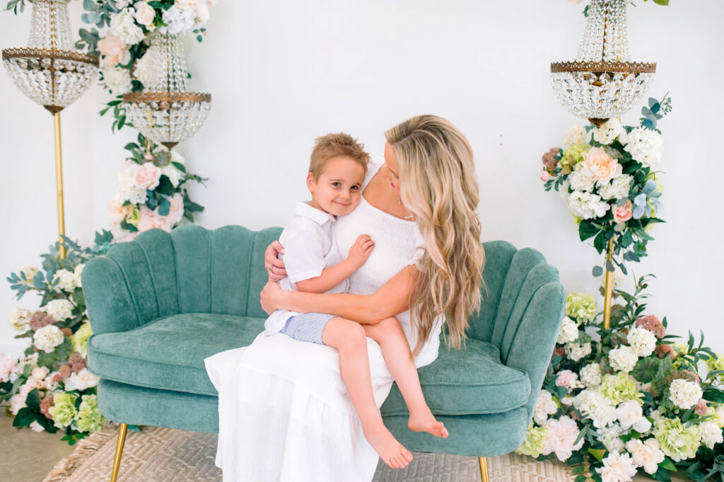 Son sitting on moms lap smiling at the camera sitting on a green couch during a mommy and me portrait session at Lemondrop Studios in Mckinney, Texas. 