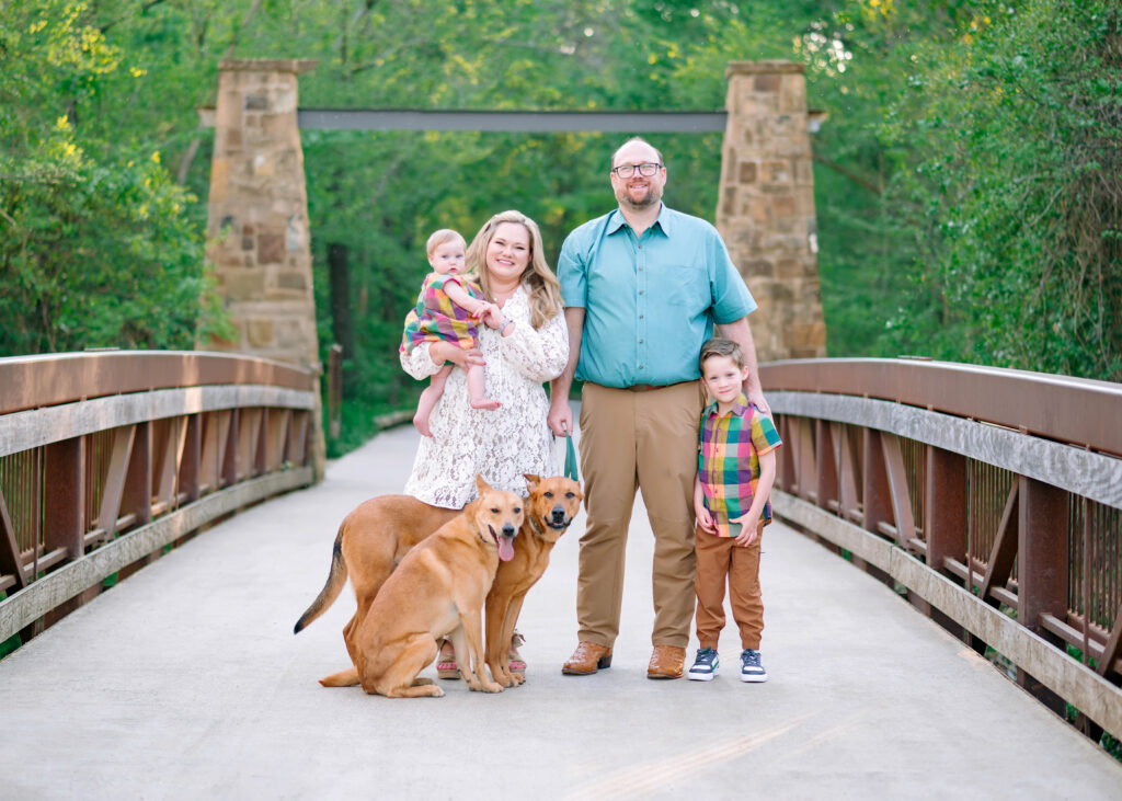 Family with 2 kids and 2 dogs standing and smiling on bridge in Plano, Texas during their family session with Jennifer L Kirk Photography. 