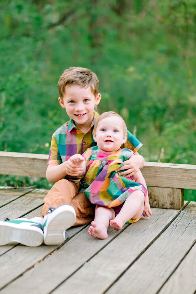 Big brother holding baby sister sitting on wooden path during their family session with Jennifer L Kirk Photography in Plano, Texas. 